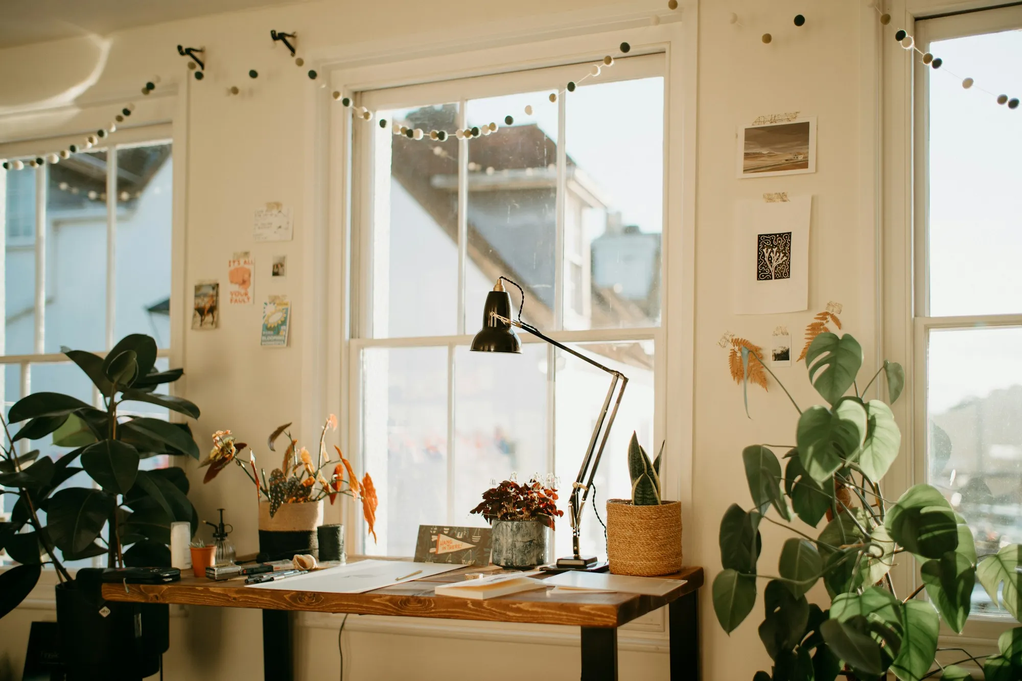 Escritorio de trabajo junto a una ventana, con plantas, lámpara y papeles, bañado en luz cálida.
