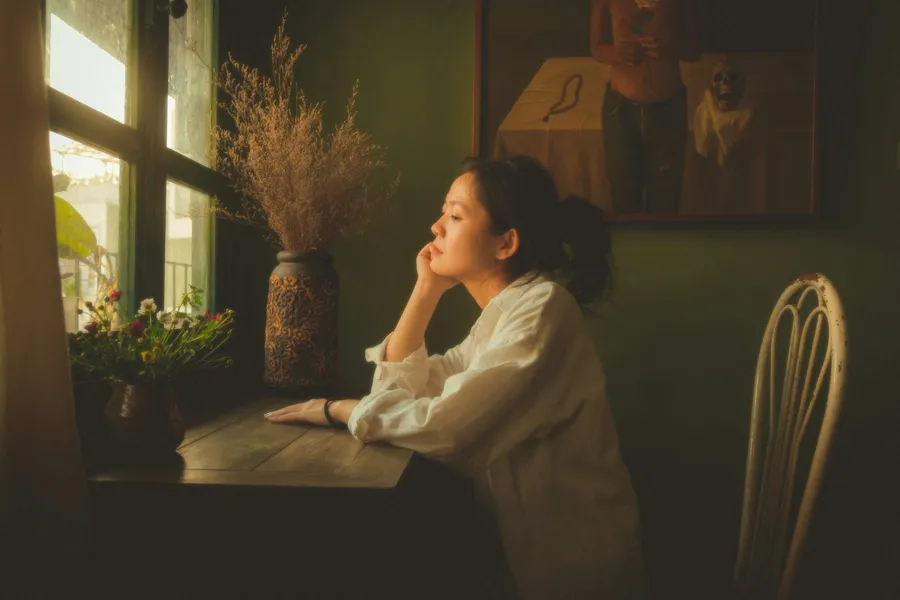 Una persona sentada junto a la ventana, pensando, con luz cálida de tarde.